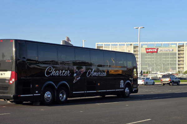 Rear-side view of a black Charter America motorcoach parked in a spacious parking lot, with Levi's Stadium visible in the background under a clear sky.