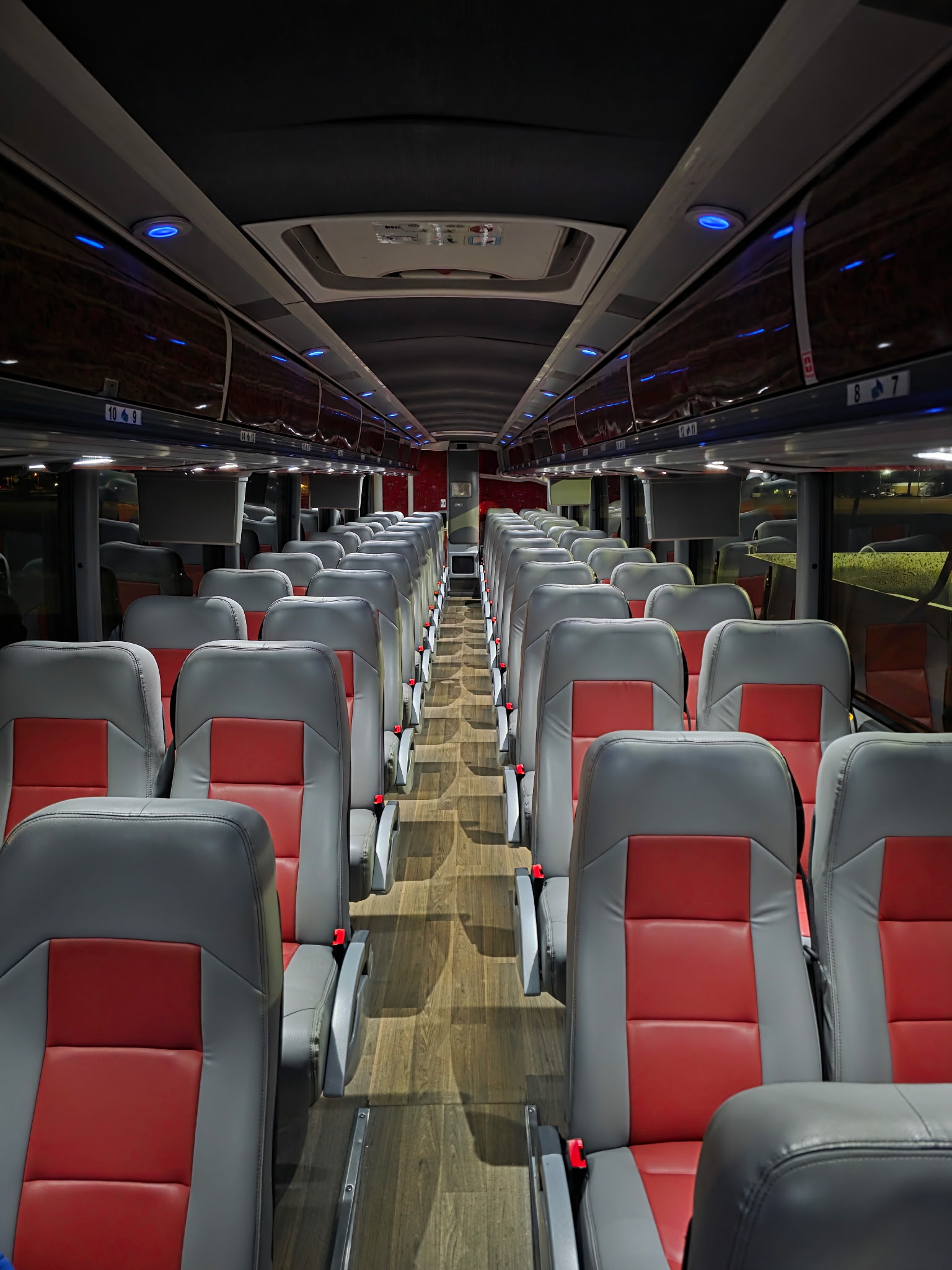 Interior view looking down the center aisle of an empty Charter America motorcoach, showing modern gray leather seats with red center inserts, wood-plank flooring, overhead luggage racks with blue accent lighting, and small video screens mounted along the ceiling.