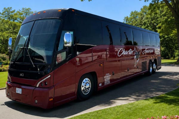 wedding-bus Profile view of a sleek, black Charter America luxury charter bus parked next to a manicured green lawn, with tall pine trees and mountains visible under a clear blue sky.