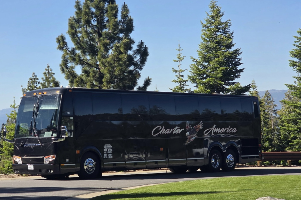 Profile view of a sleek, black Charter America luxury charter bus parked next to a manicured green lawn, with tall pine trees and mountains visible under a clear blue sky.